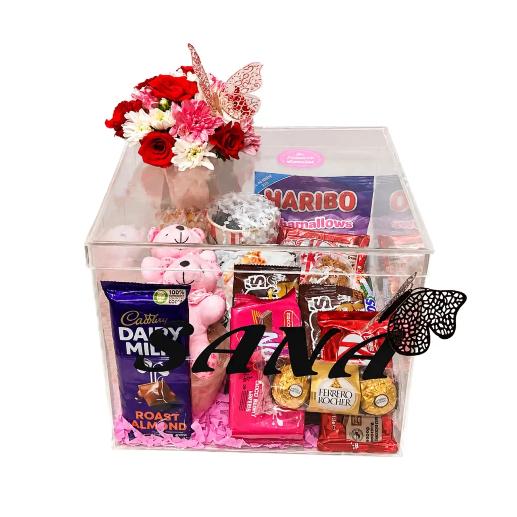 A clear box filled with assorted treats and colorful flowers, showcasing a delightful gift arrangement.
